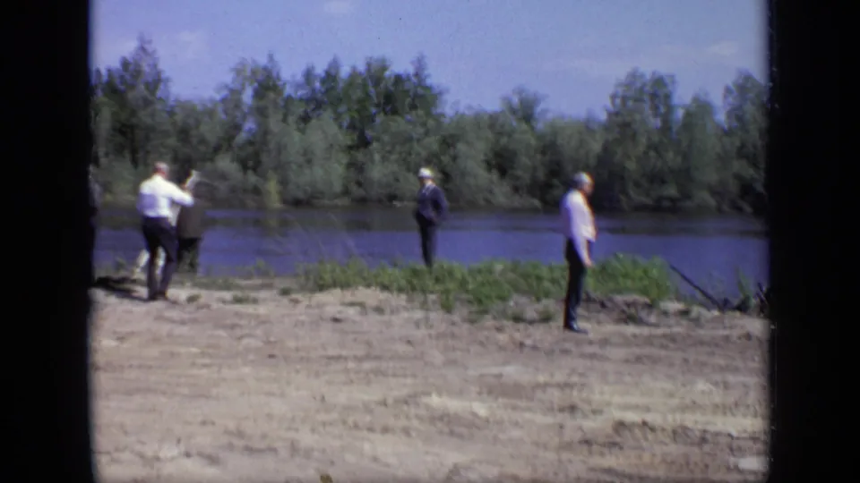 1971 Moscow home-movie frame of men gathered by the riverbank, with one raising a camera or arm toward another.