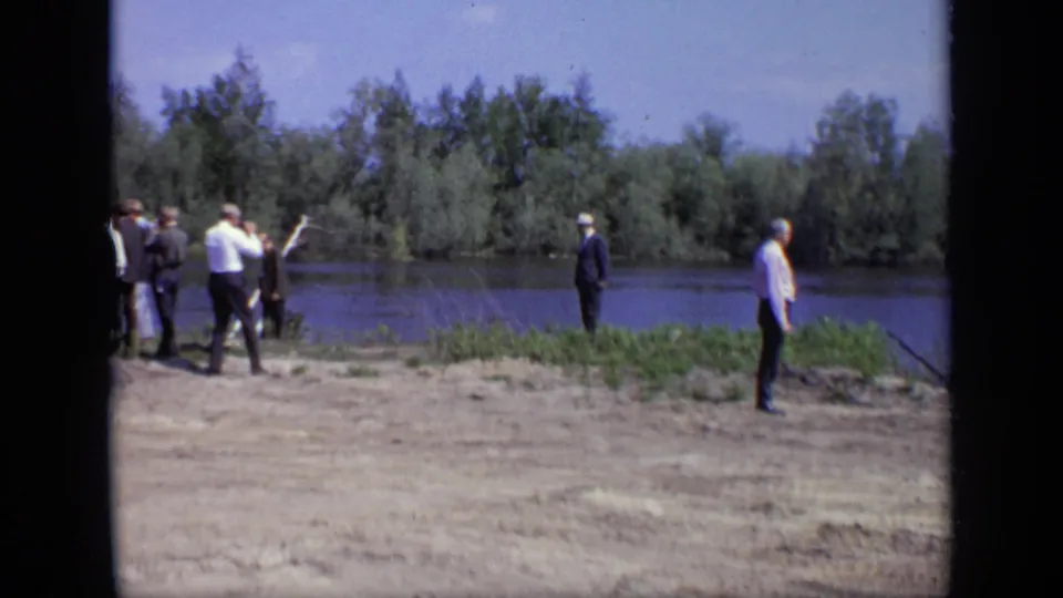 1971 Moscow frame showing a group of adult men on a dirt riverbank with calm water and dense trees beyond.