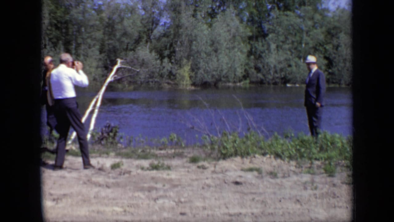 1971 Moscow home-movie frame of adults on a sandy riverbank, including a man with binoculars and a suited man in a light hat.