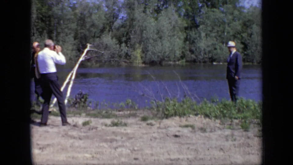 1971 Moscow home-movie frame of adults on a sandy riverbank, including a man with binoculars and a suited man in a light hat.