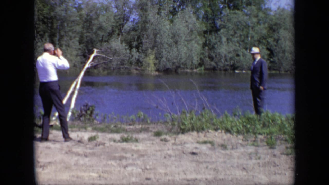 A man looks through binoculars toward the river while a suited man in a light hat stands nearby.