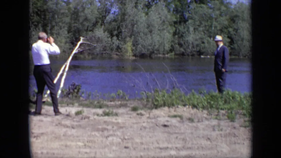 1971 Moscow home-movie view of two men on a riverbank, with binoculars, a pale branch or pole, water, and trees behind them.