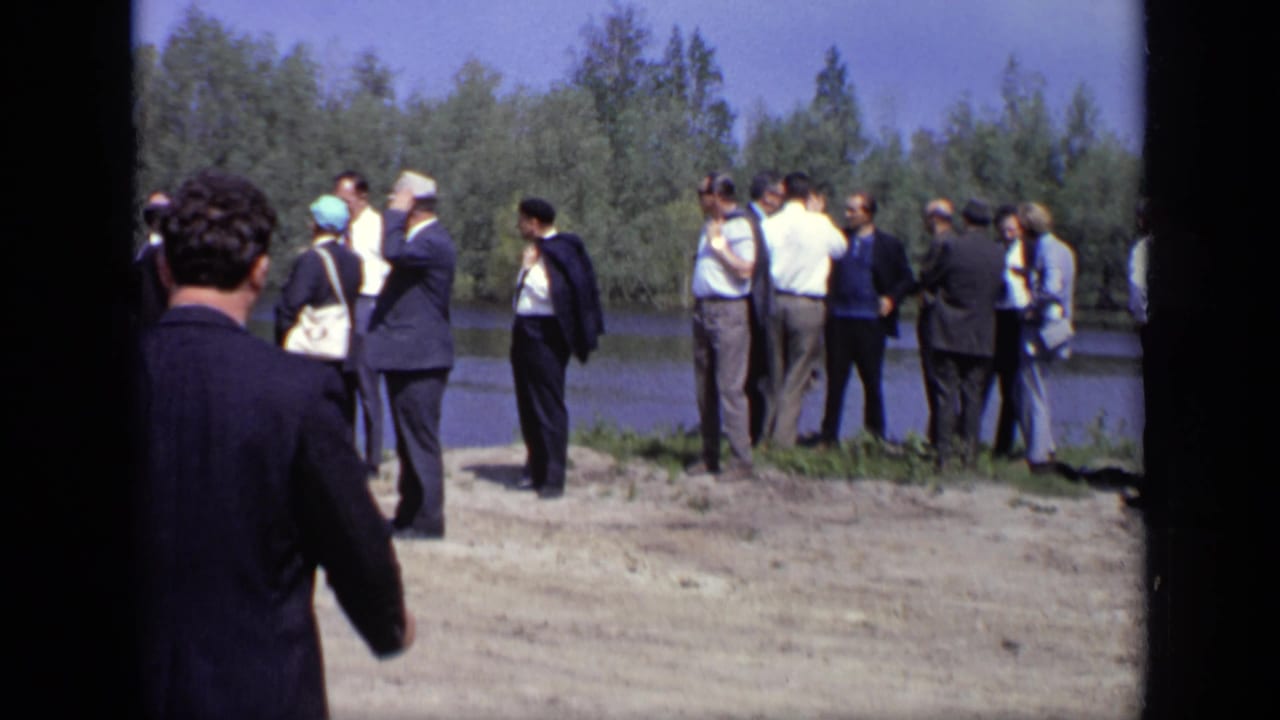 In 1971 Moscow footage, adults in suits and hats stand in small groups on a sunlit riverbank.