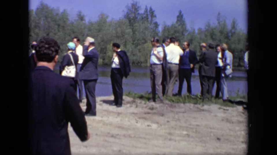 In 1971 Moscow footage, adults in suits and hats stand in small groups on a sunlit riverbank.