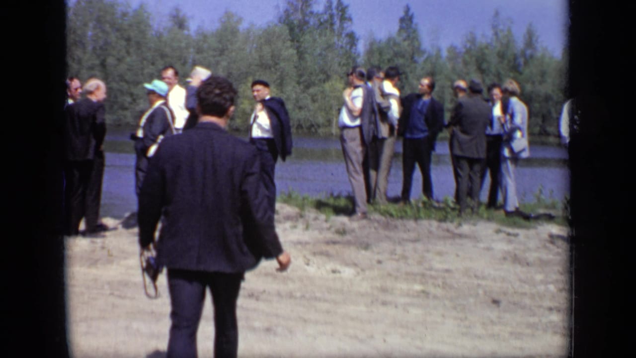 A man in a dark suit walks toward a group of adults beside the water in 1971 Moscow home-movie footage.