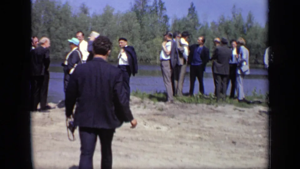 A man in a dark suit walks toward a group of adults beside the water in 1971 Moscow home-movie footage.