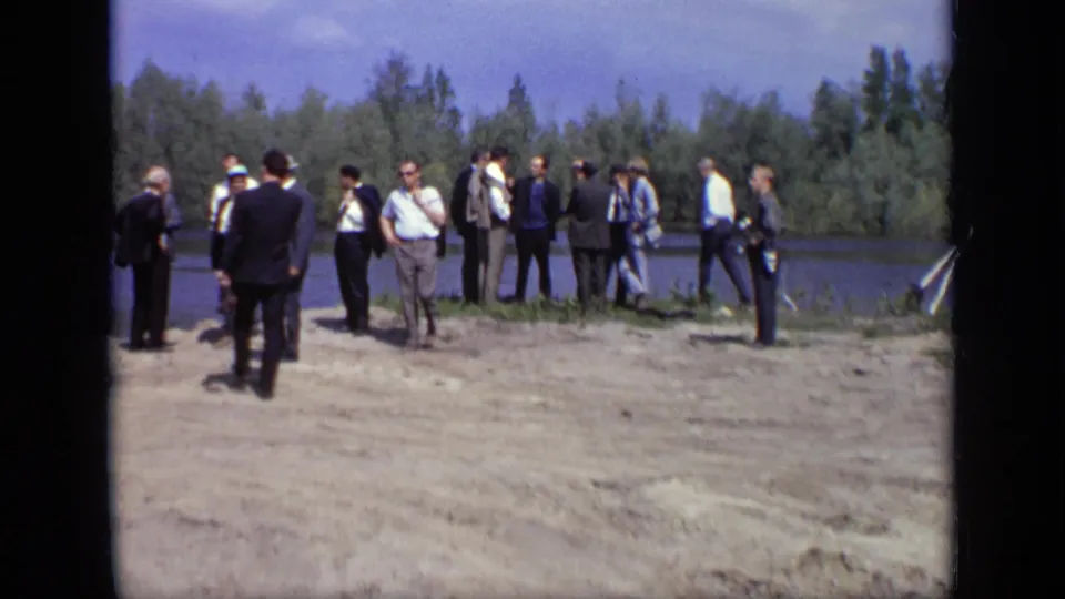 Adults in formal and casual clothing stand along a Moscow riverbank, with water and trees in the background.