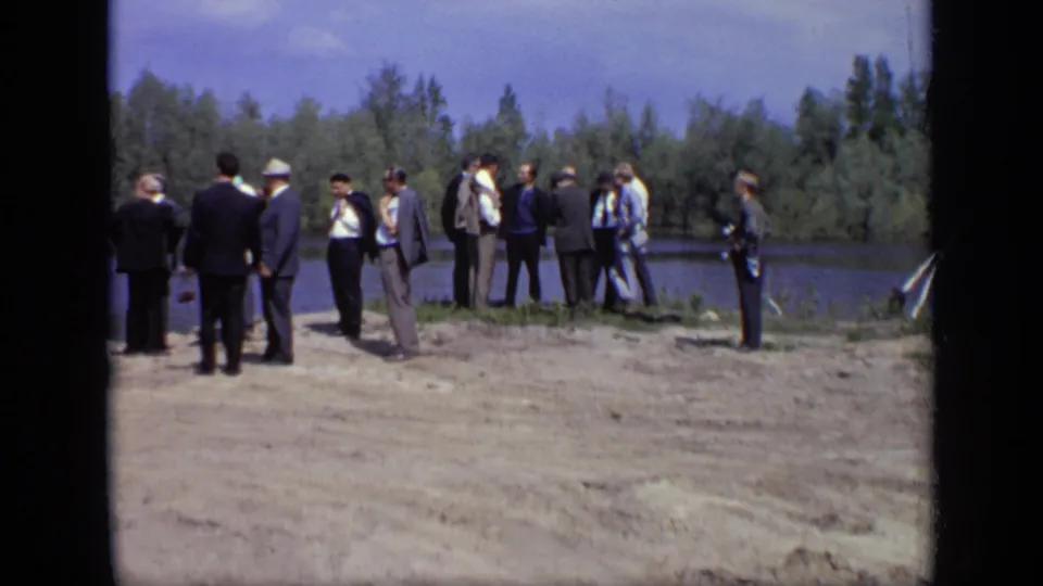 A small group of adults converse near the river in 1971 Moscow, framed by trees and sandy ground.