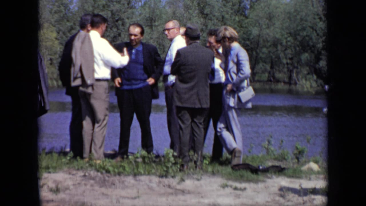 Men in suits stand and talk on a grassy bank beside a lake.