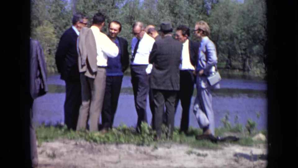 Adult men in suits and light shirts stand near a Moscow lakeside, with water and trees visible in the background.