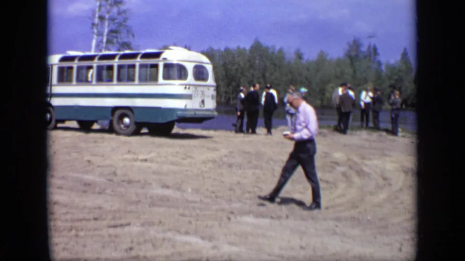 A lone adult man crosses an open dirt area while a white and blue bus and distant group stand near the lake.