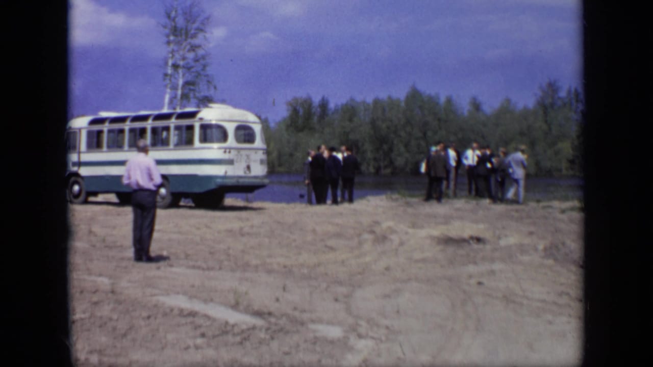 Men stand near a white and teal bus on a dirt area beside a river and trees.