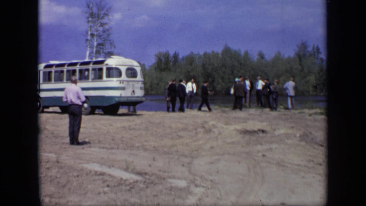 Men in suits and casual shirts stand near a parked bus by a river in the supplied 1971 Moscow home-movie clip.