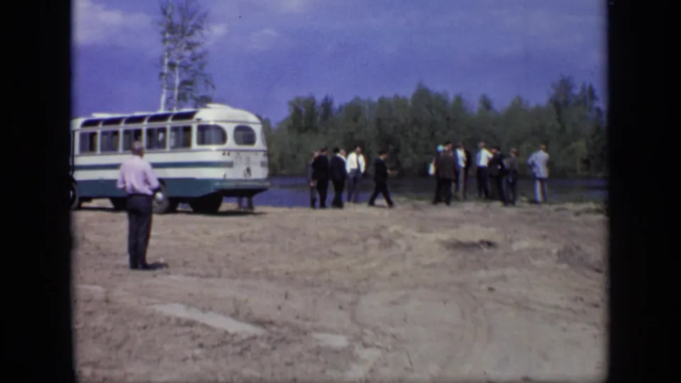 Men in suits and casual shirts stand near a parked bus by a river in the supplied 1971 Moscow home-movie clip.