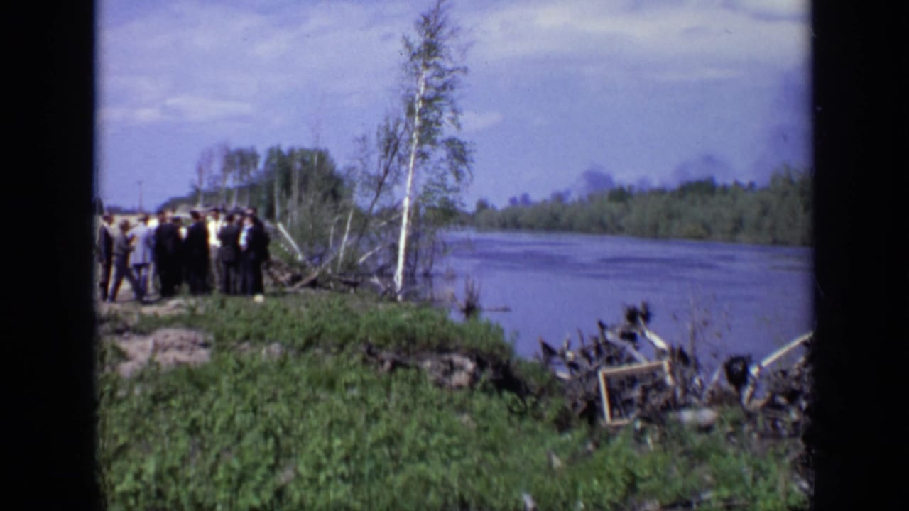 Men gathered on a grassy riverbank beside trees and shoreline debris.