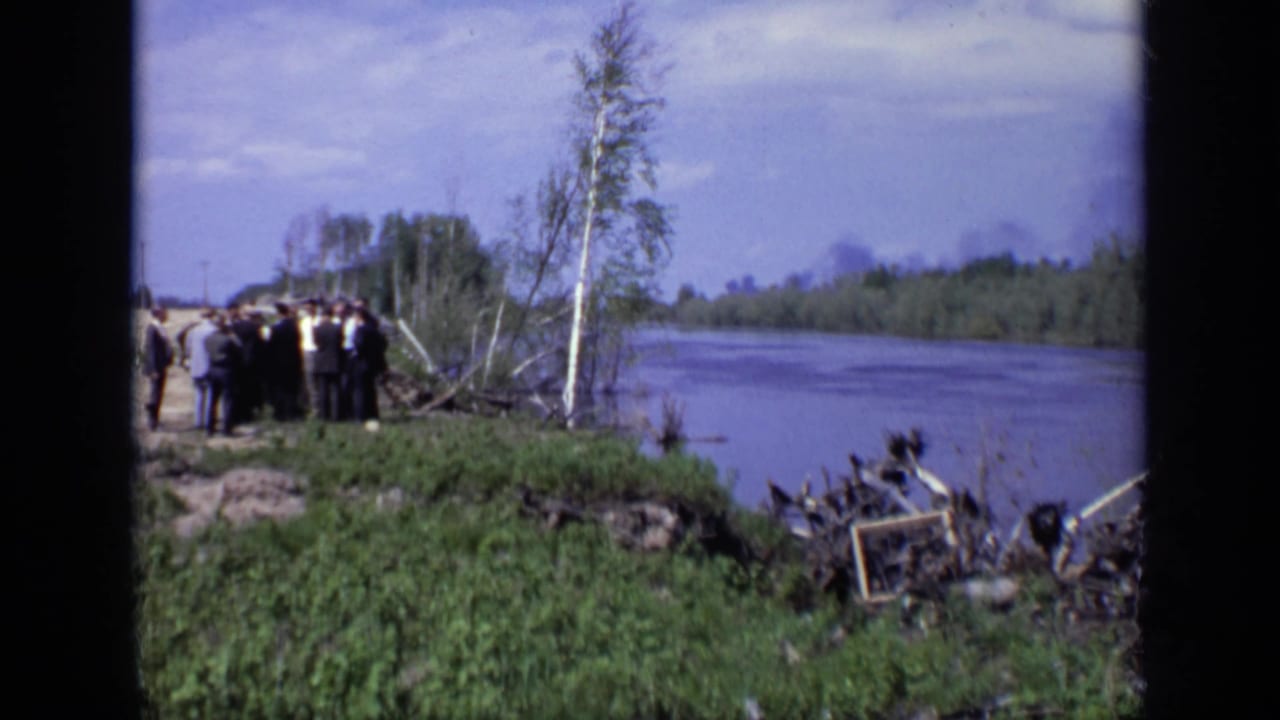 Distant 1971 Moscow riverside view with suited men gathered near the water, a thin tree in front, and debris scattered at the shoreline.