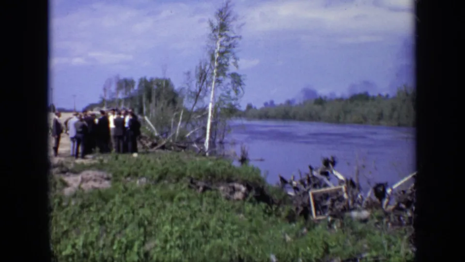 Distant 1971 Moscow riverside view with suited men gathered near the water, a thin tree in front, and debris scattered at the shoreline.