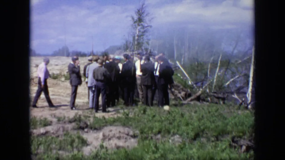 1971 Moscow 8mm frame showing suited men on a dirt roadside beside a riverbank, with haze, trees, and shoreline debris nearby.