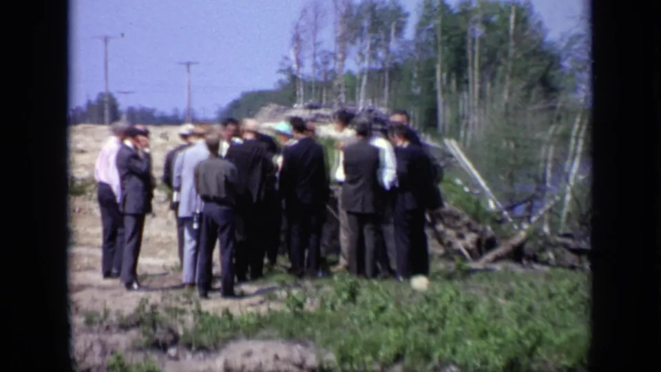 1971 Moscow riverside frame with a group of men in suits and hats on a dirt road beside trees, utility poles, grass, and water.