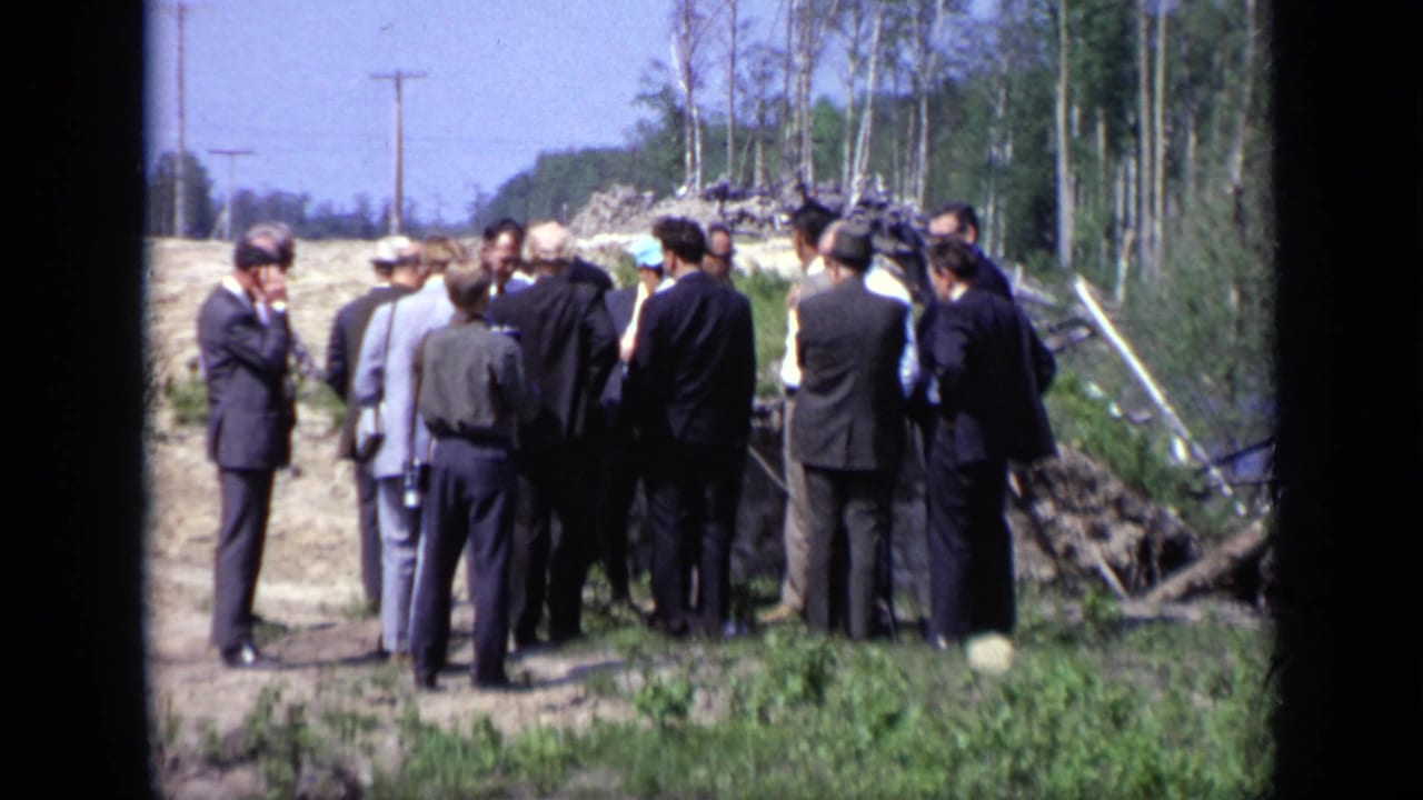 Suited men gather on a dirt riverbank near trees and utility poles.