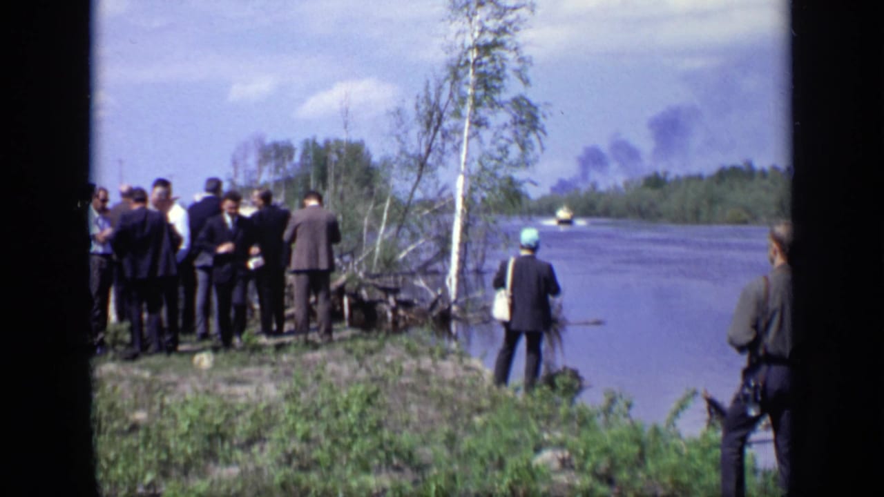 Group of men stands beside a river while smoke rises across the water.