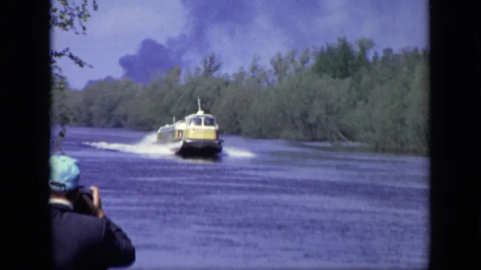 A yellow speedboat moves toward the camera on a Moscow river, with wooded banks, visible wake, and a spectator at the lower left.