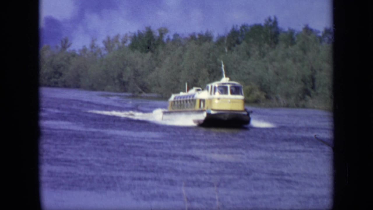 The yellow hydrofoil continues across the lake near Moscow, with its wake spreading behind it and trees in the background.