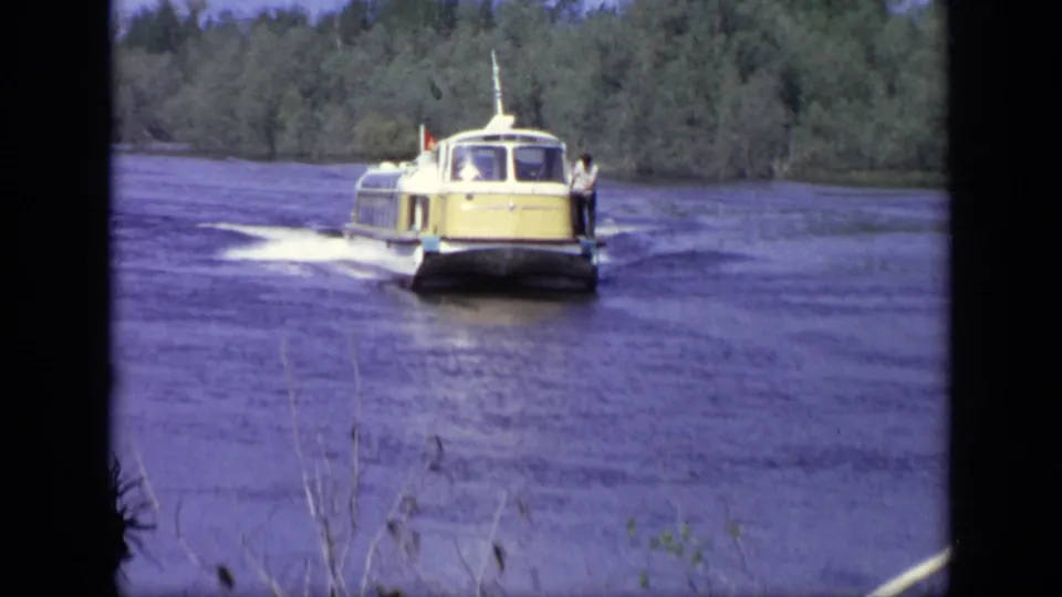 The yellow hydrofoil faces nearer to camera on the lake, with a small person visible along its side and trees behind.