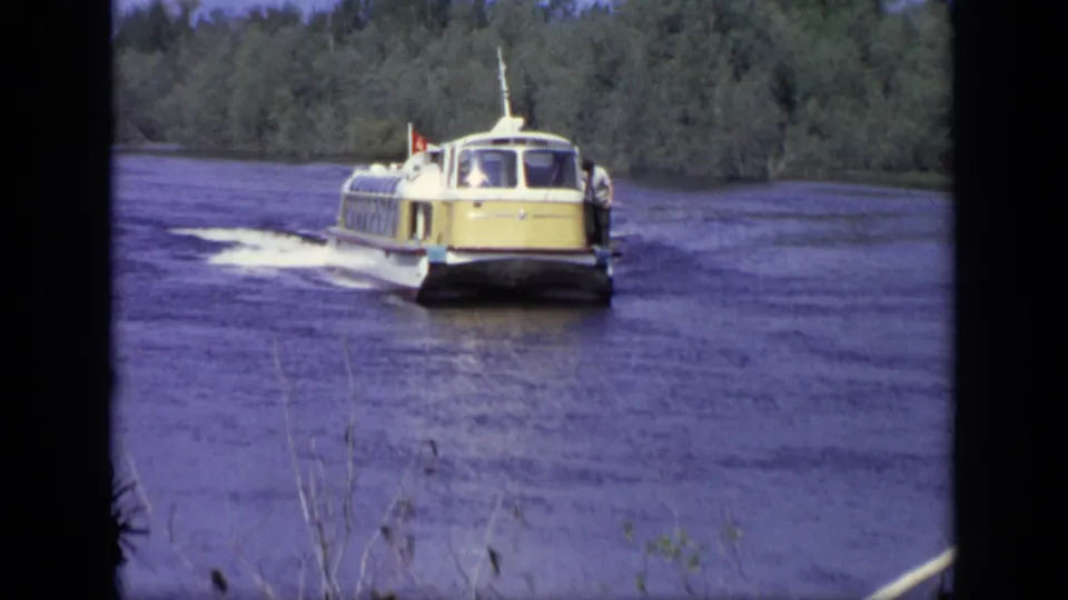 1971 Moscow home-movie frame showing a yellow-and-white passenger boat approaching on a wide river with trees behind it.