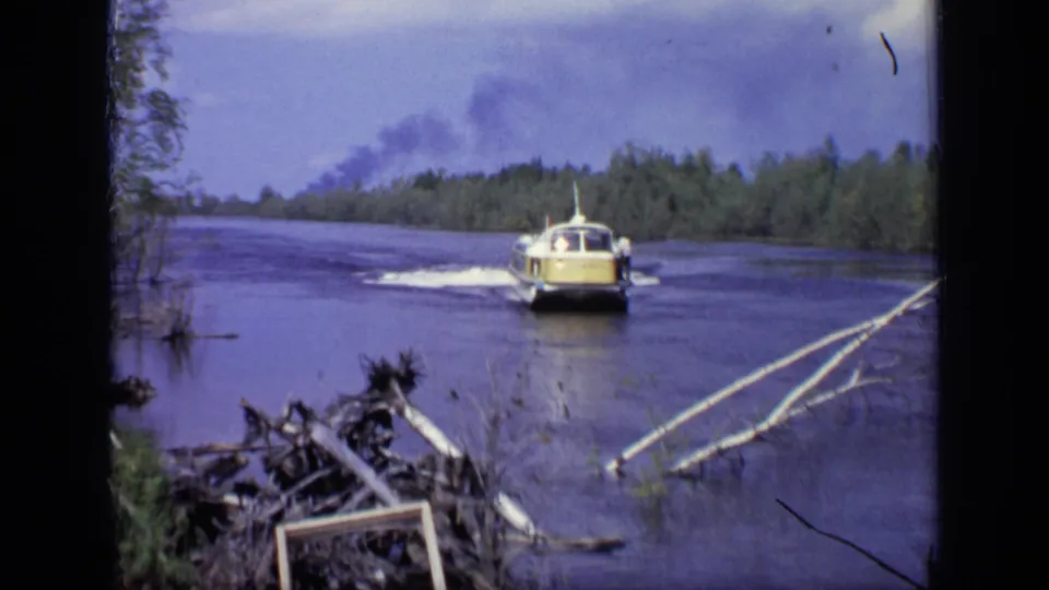 1971 Moscow home-movie frame showing the passenger boat moving away on a river, with dark smoke rising beyond the wooded bank.
