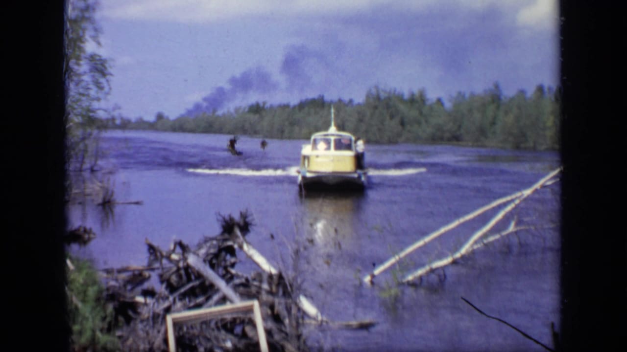 1971 Moscow home-movie frame showing a yellow and white hovercraft moving toward a wooded shore with wake behind it.