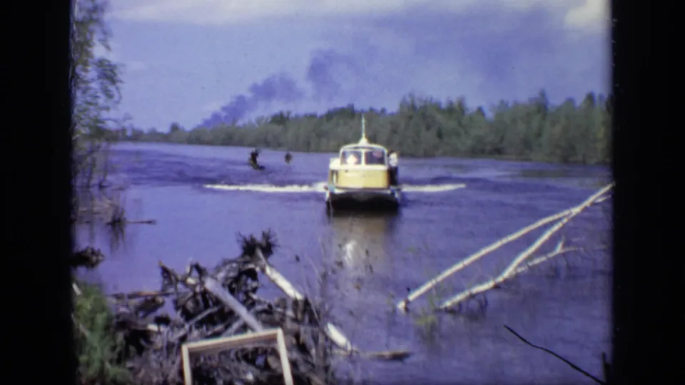1971 Moscow home-movie frame showing a yellow and white hovercraft moving toward a wooded shore with wake behind it.