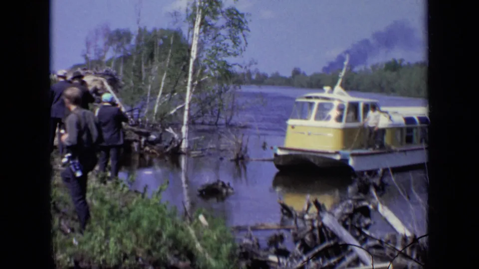 1971 Moscow home-movie frame showing several people on a grassy wooded bank beside the arriving yellow and white hovercraft.