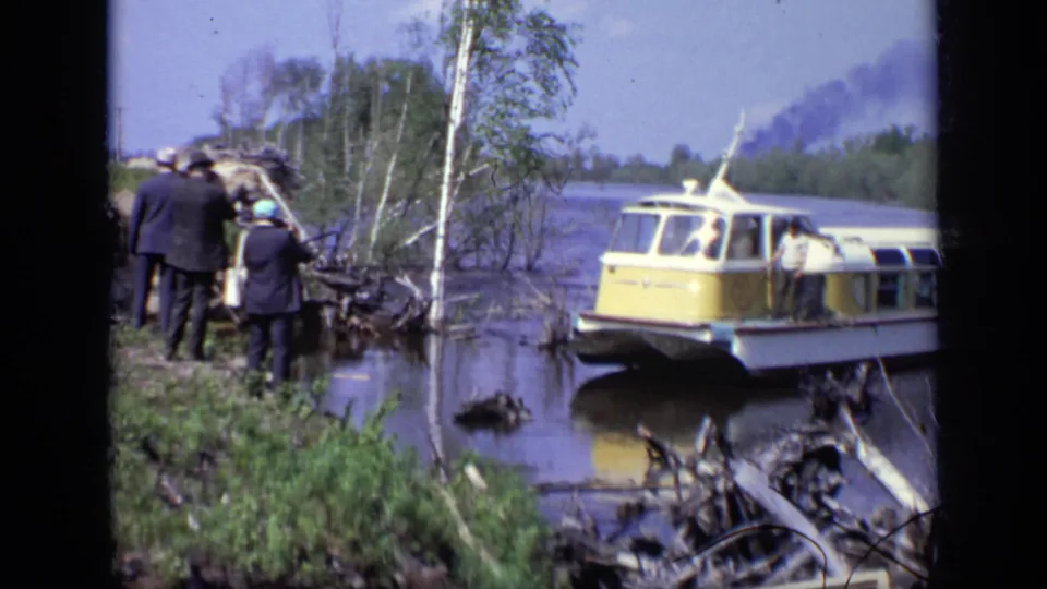 1971 Moscow frame showing people in dark clothing near the wooded bank as the yellow and white hovercraft rests close to shore.