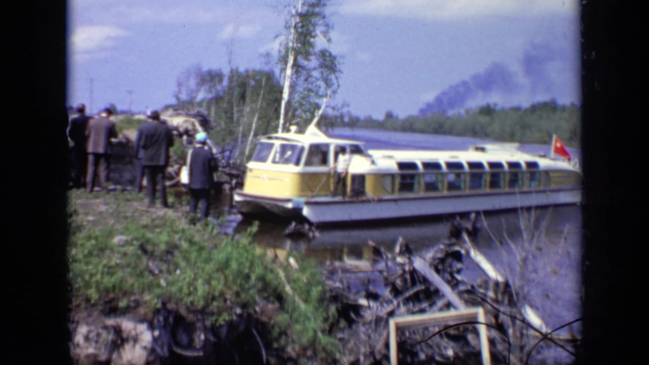 1971 Moscow frame with a long yellow-and-white passenger boat at the bank, adults in dark clothing nearby, and smoke beyond the treeline.