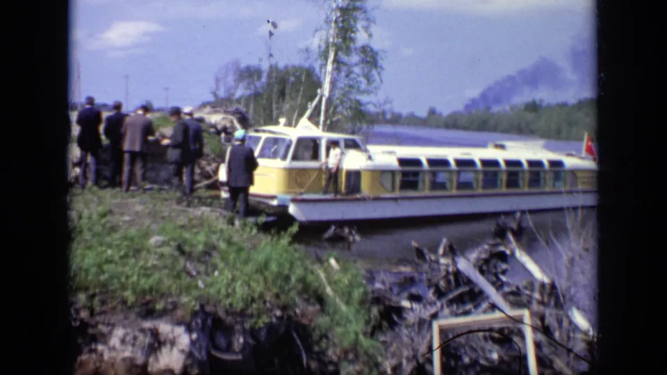 1971 Moscow riverbank view of a yellow-and-white passenger boat with suited adults standing nearby and dark smoke in the distance.