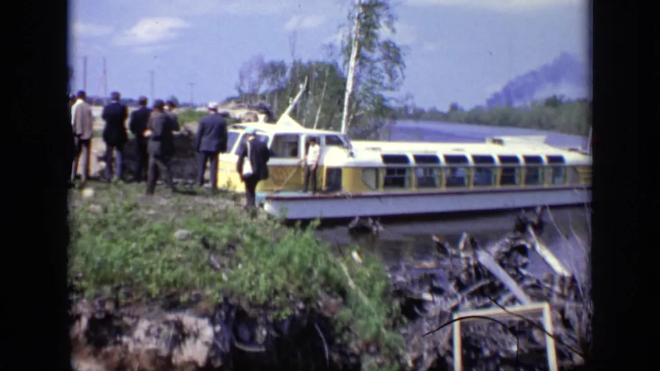 1971 Moscow home-movie frame showing adults beside a yellow-and-white river boat, with shoreline debris and a smoky background.