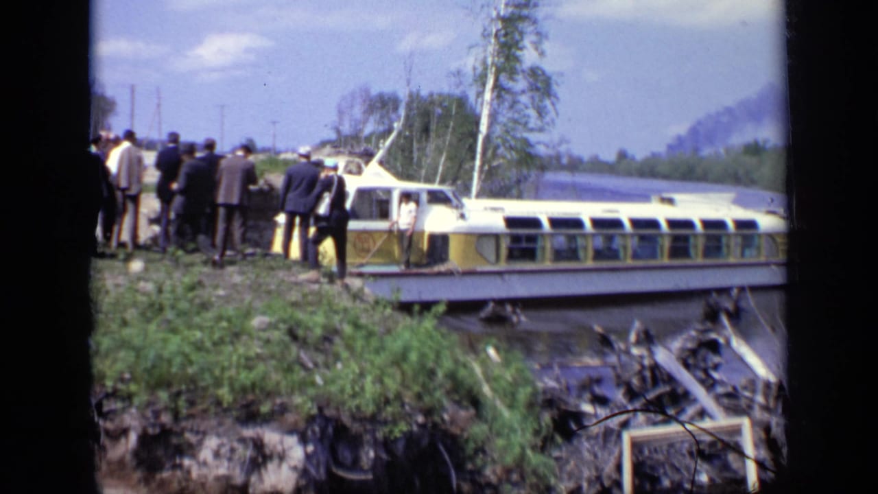 Men in suits stand on a grassy riverbank beside a yellow and white ferry.