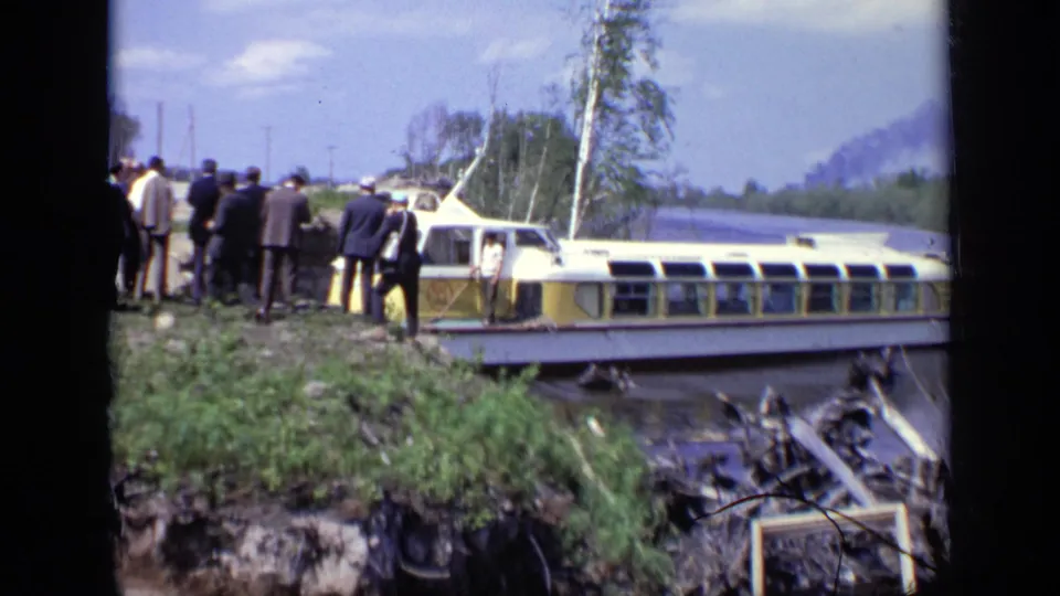 1971 Moscow home-movie frame of suited men gathered near a yellow and white ferry at a grassy riverbank.