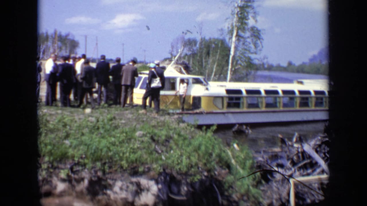 A group of suited men stands near the ferry landing on a grassy bank.