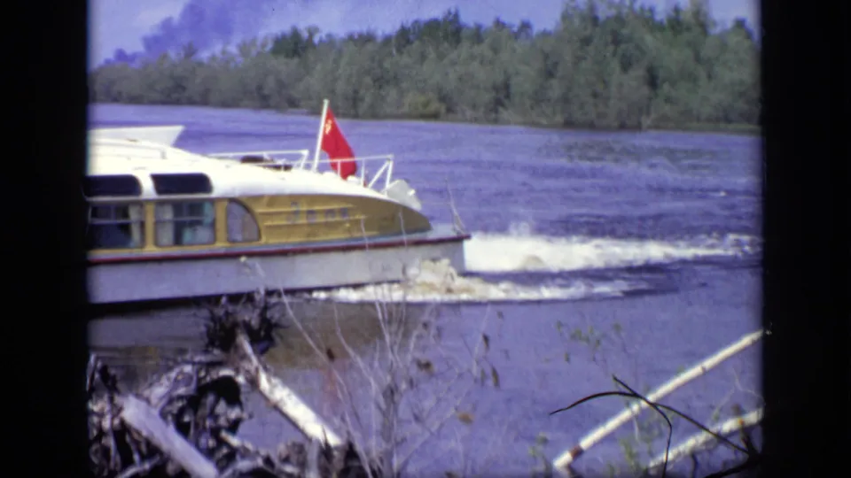 A yellow and white river ferry with a red flag moves past the wooded riverbank in 1971 Moscow footage.