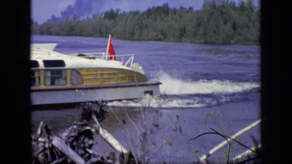 1971 Moscow 8mm frame of a yellow and white motorboat passing along a river, leaving a foamy wake near the bank.