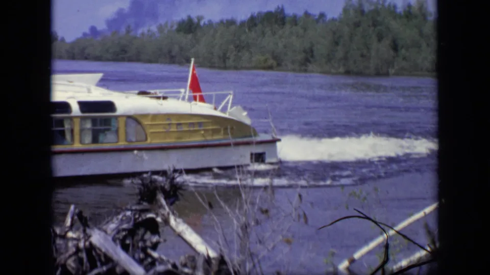 The small river boat continues past the camera in a handheld 1971 Moscow home-movie shot, with trees beyond the water.