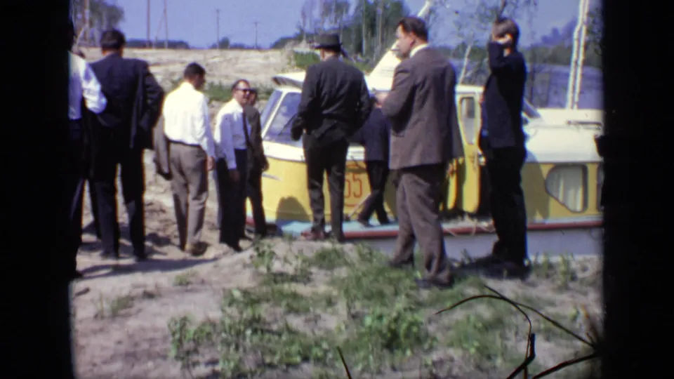 Men in suits and light shirts gather by a yellow and white tour boat on a grassy Moscow riverbank in 1971 home-movie footage.