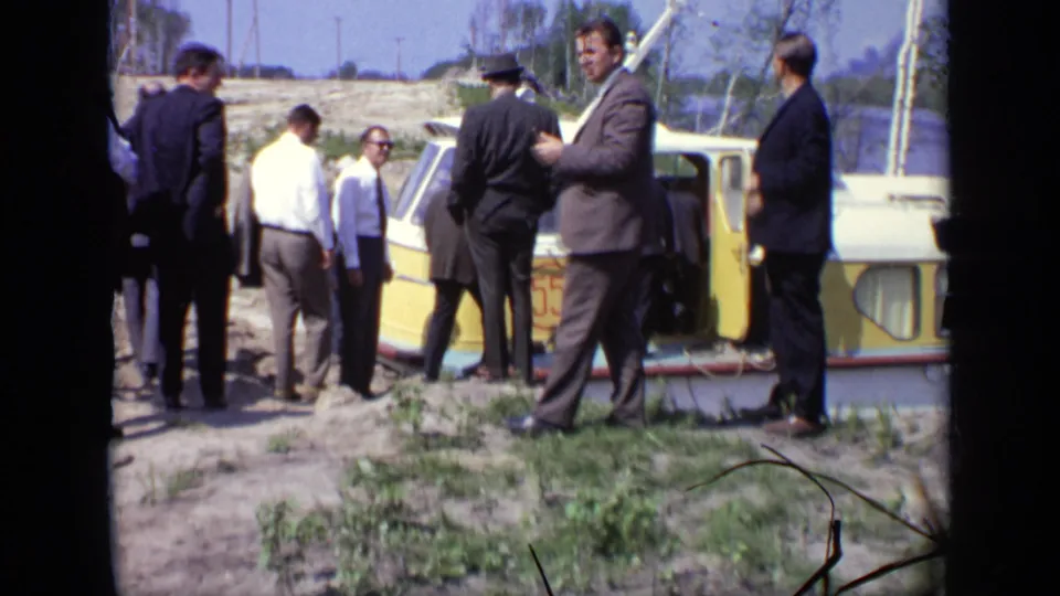 Adult men in business attire and casual shirts stand beside the boat during boarding from a rough grassy riverbank in Moscow, 1971.
