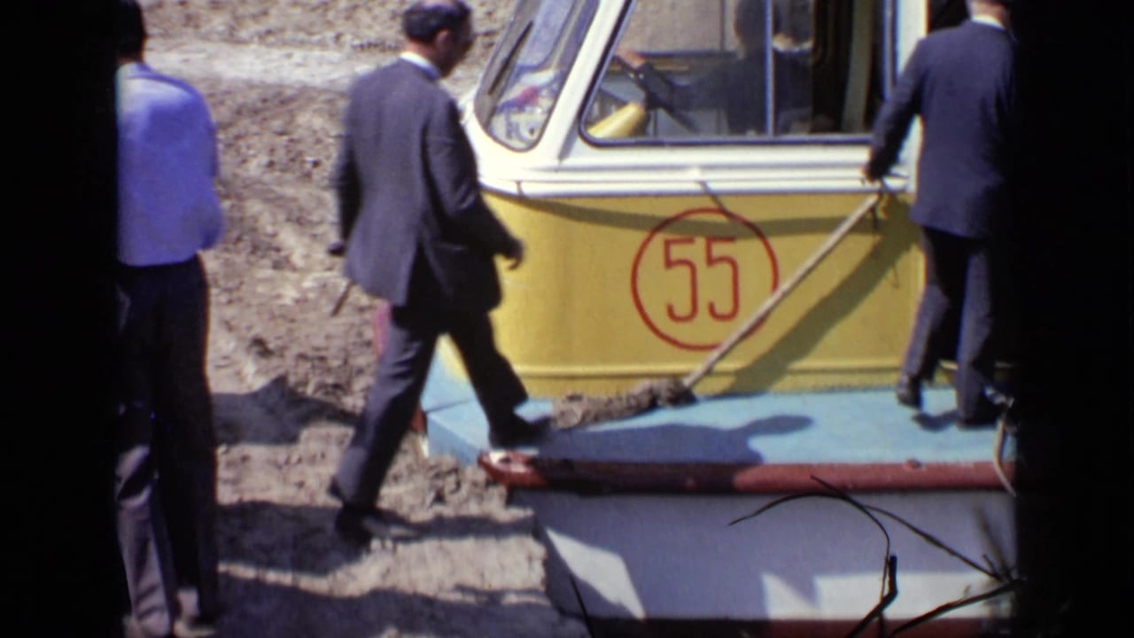 Men in suits walk beside a yellow and blue boat marked 55.