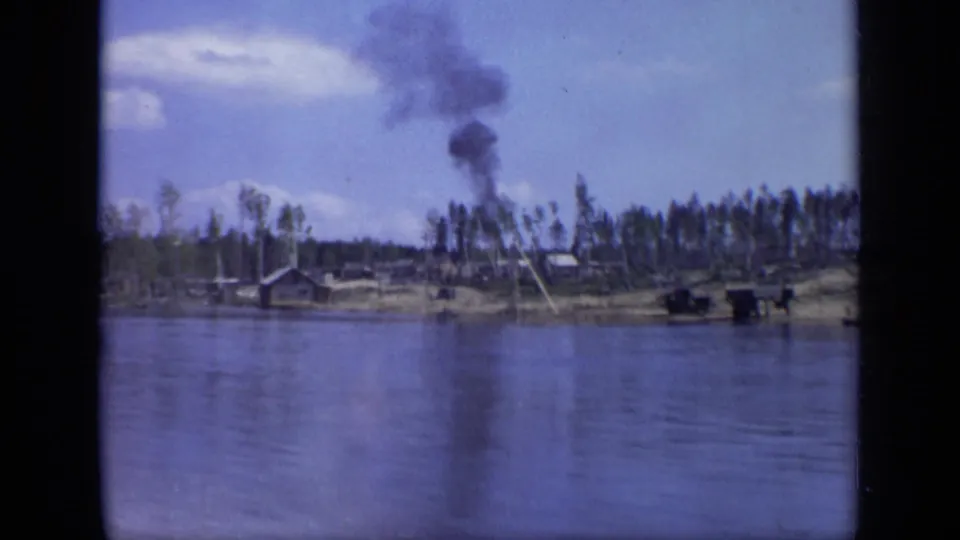 1971 Moscow home-movie frame looking across the river toward simple buildings and smoke behind the tree line.