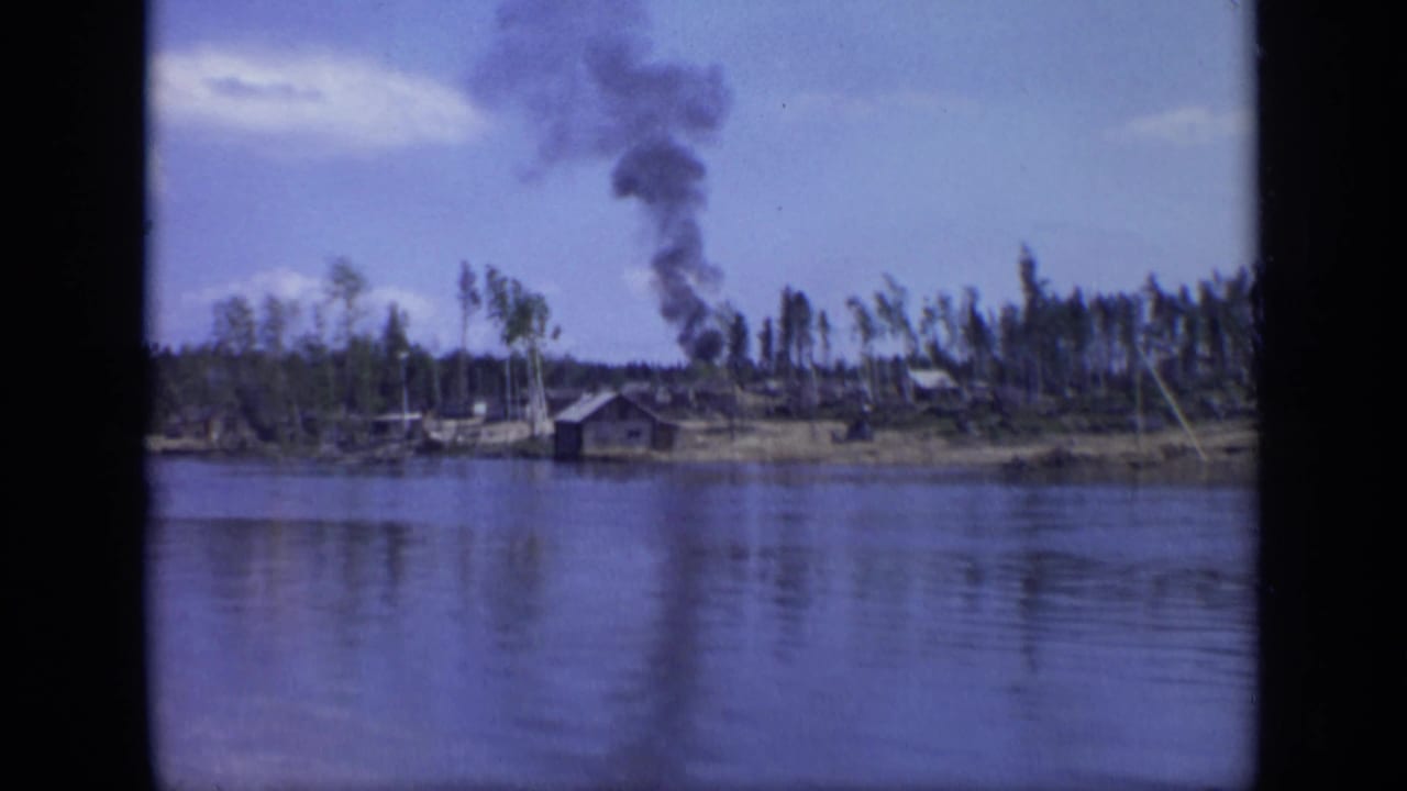 1971 Moscow 8mm frame showing dark smoke above a low riverside industrial area with wooden structures and sparse trees.
