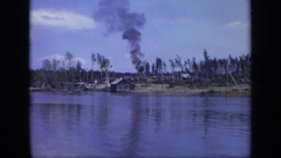 1971 Moscow 8mm frame showing dark smoke above a low riverside industrial area with wooden structures and sparse trees.