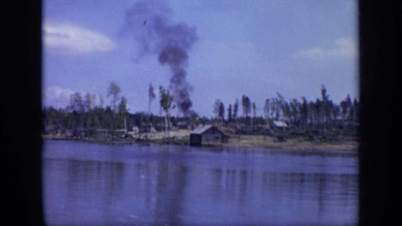 1971 Moscow-area river view with a dark industrial smoke plume rising behind a small cabin and sparse trees.